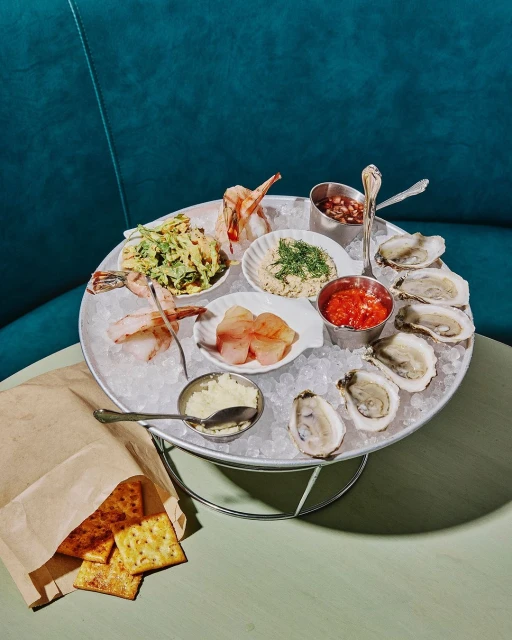 Overhead shot of a seafood platter with oysters, shrimp, sauces, and crackers, displayed on ice on a light green table.