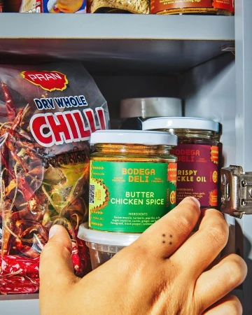 Jars of butter chicken spice and crispy pickle oil are displayed in a pantry with a bag of whole dried chilis and a hand reaching.