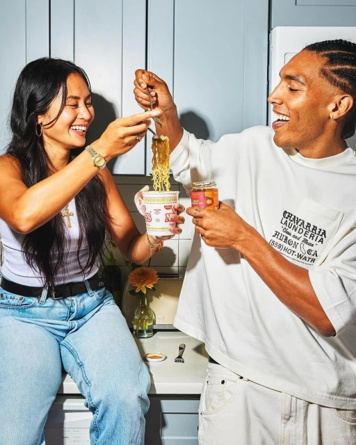A smiling man and woman are eating noodles indoors near a blue wall and a white refrigerator in a modern kitchen with warm, natural light.