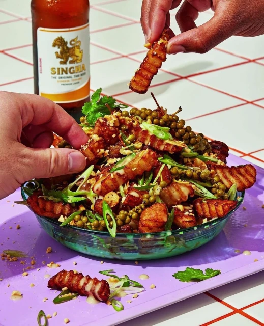 Close-up of a vibrant Thai-inspired dish with crispy pork belly, green peppercorns, herbs, and nuts served in a glass bowl with hands.