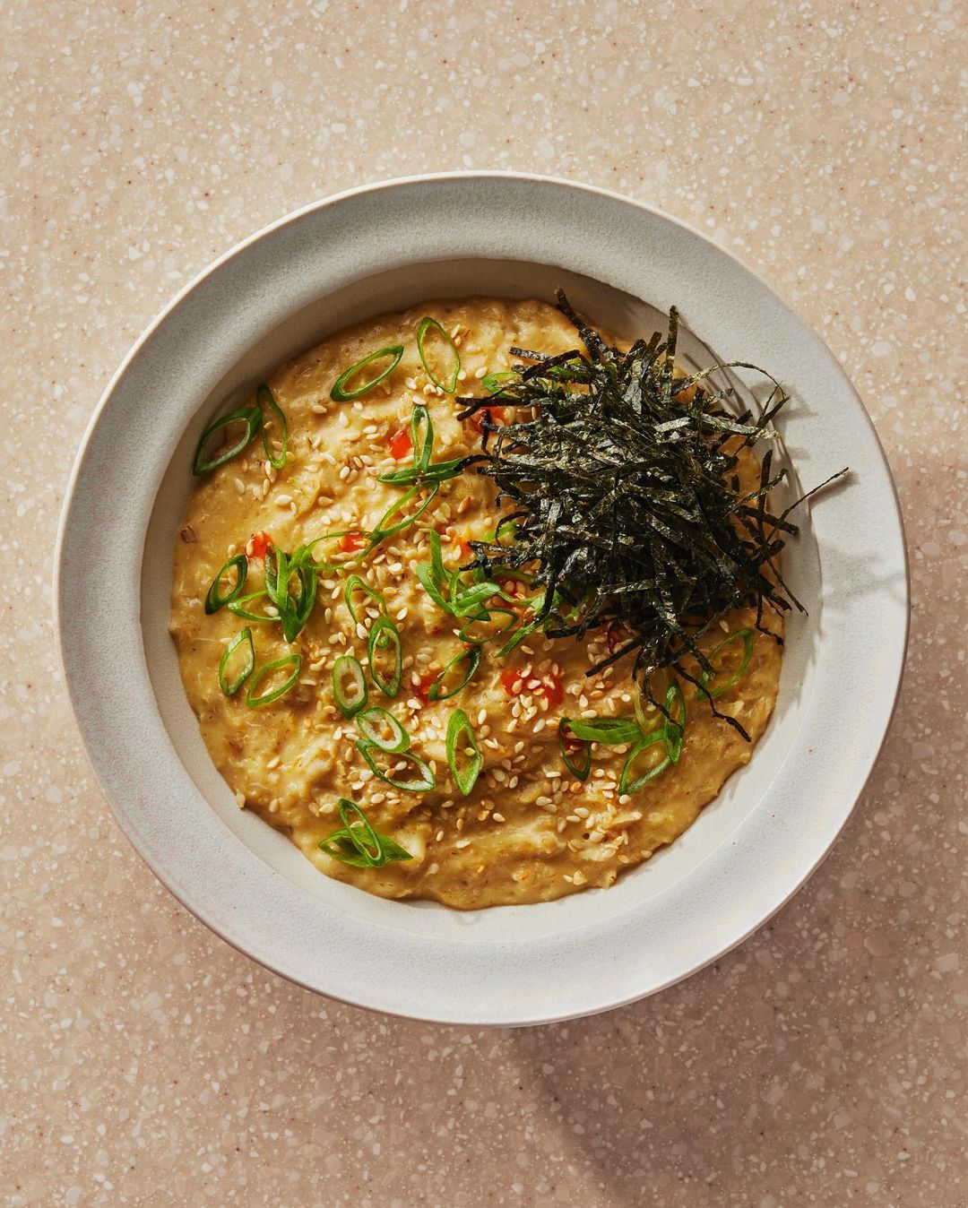 Overhead shot of a ceramic bowl filled with savoury oatmeal topped with sliced scallions, sesame seeds, and nori seaweed.