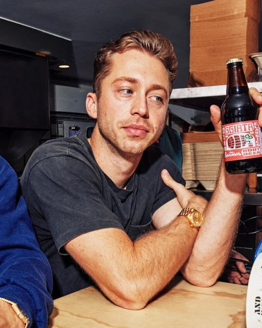 Man holding bottle of root beer with label, posing indoors at a table, wearing black t-shirt and gold watch.