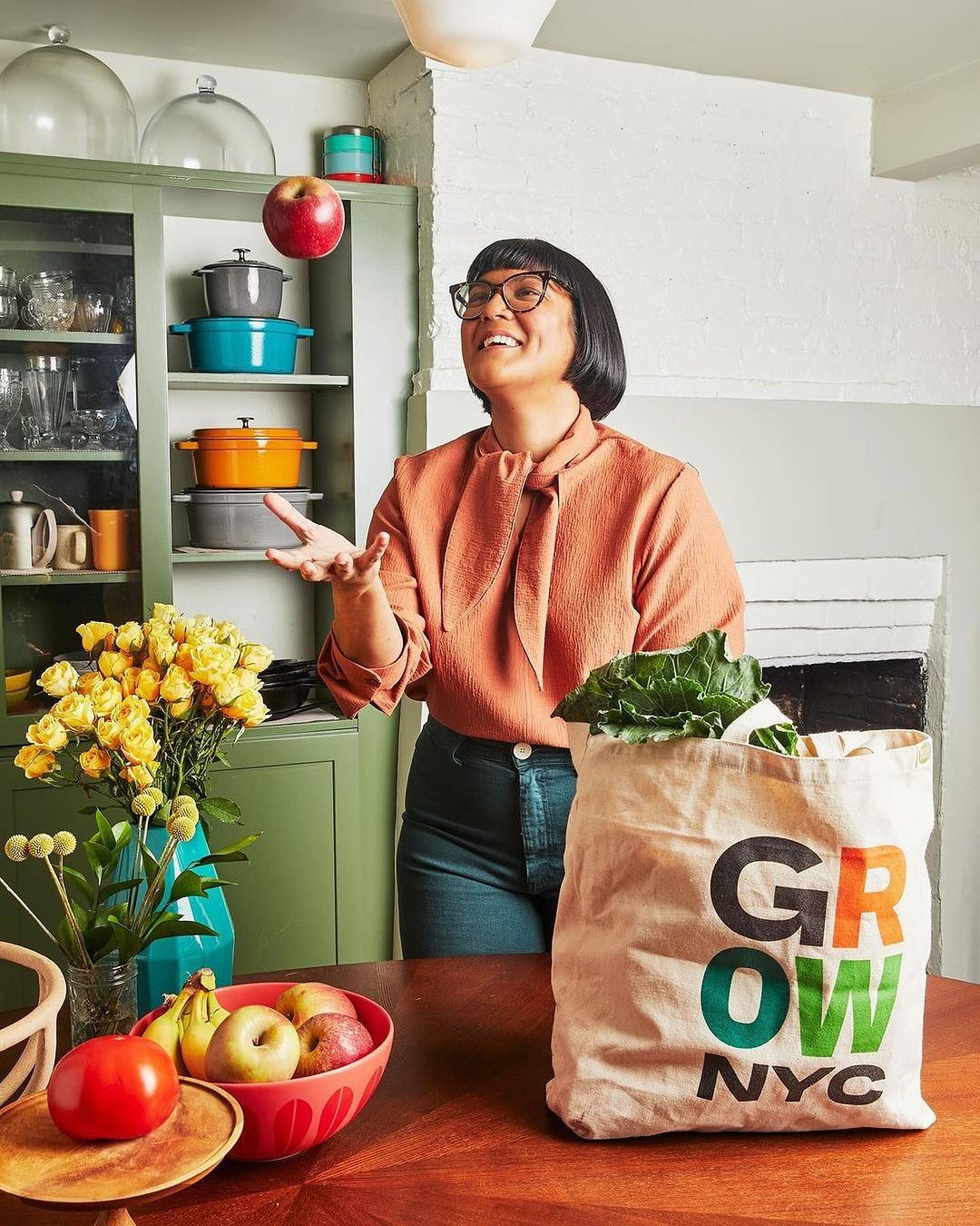 Woman in glasses juggling an apple in a kitchen with a bag of groceries and flowers on a wooden table, illuminated by overhead light.