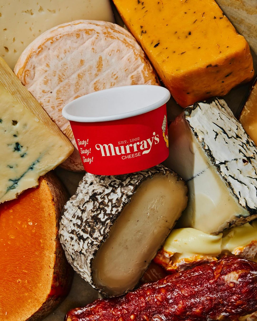 Overhead shot of assorted cheeses and cured meats with a Murray's Cheese container in a warm-toned food photography style.