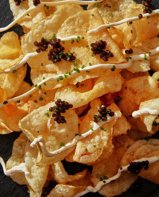 Close-up of golden potato chips topped with white sauce, black caviar, and chives, creating an indulgent food photography composition.