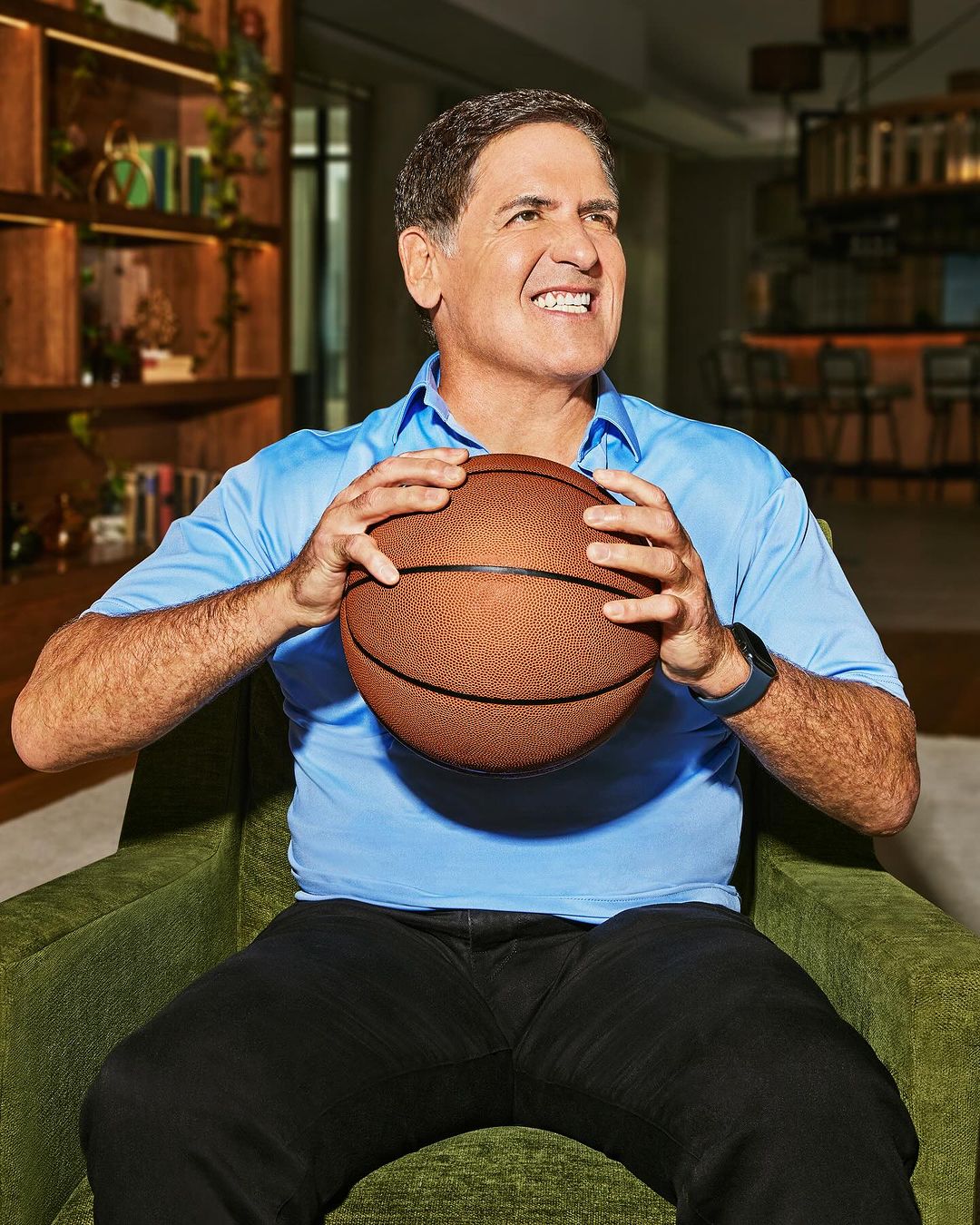 Mark Cuban in light blue shirt holds basketball while seated indoors in an arm chair, looking upward with a slightly quizzical expression.