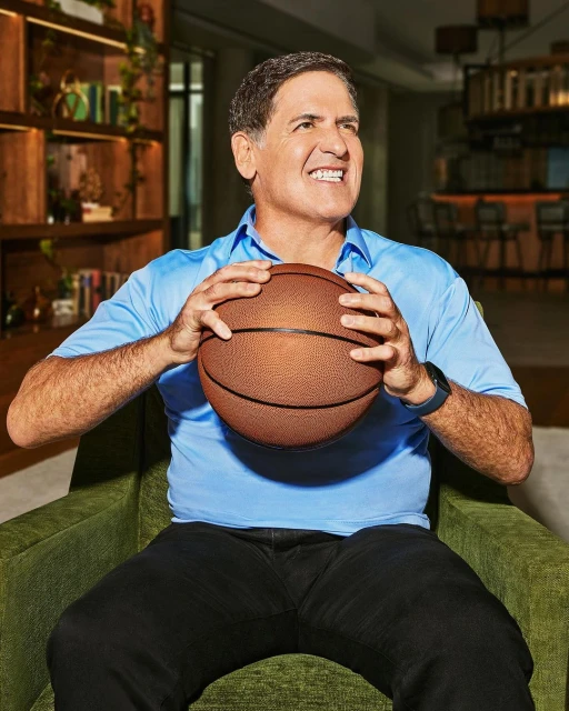 Mark Cuban in light blue shirt holds basketball while seated indoors in an arm chair, looking upward with a slightly quizzical expression.