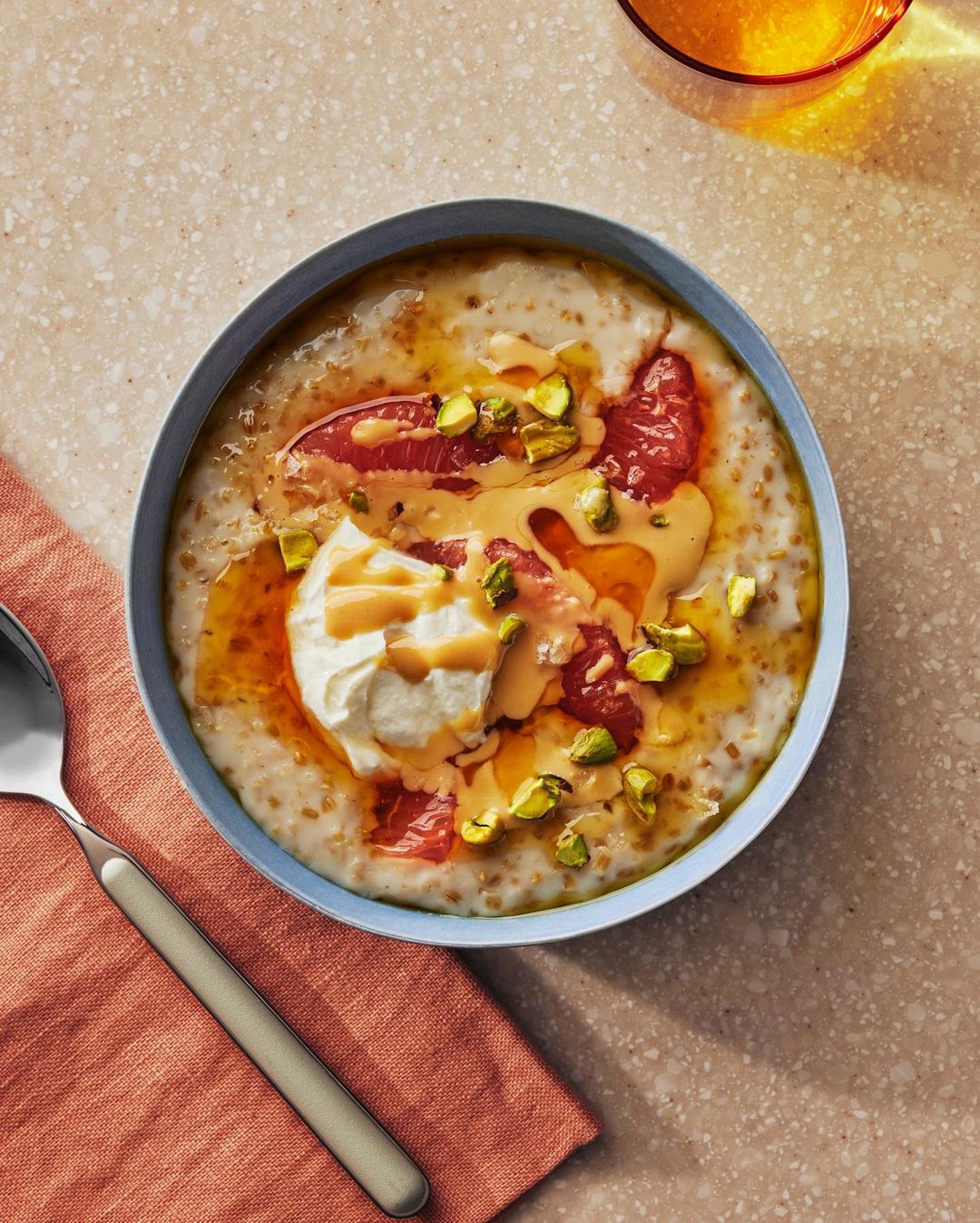 Overhead shot of a breakfast bowl with porridge, grapefruit, cream, pistachios, and honey, accompanied by a spoon and coral-colored napkin.