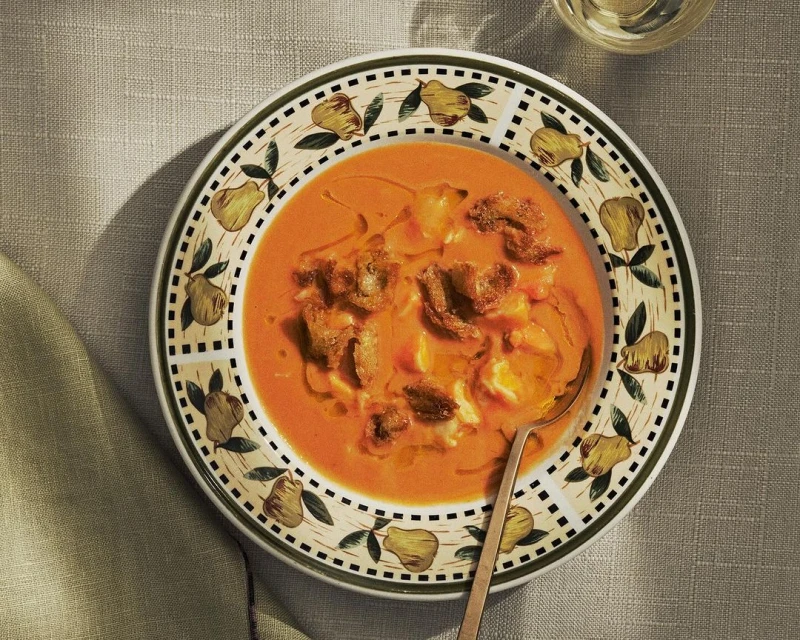 Overhead shot of orange soup with croutons, white cheese, and a spoon on a decorative plate, sitting on a linen tablecloth.