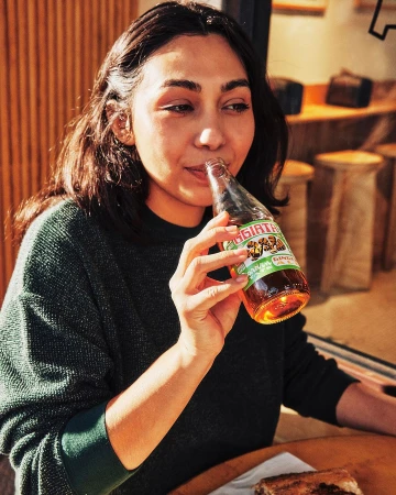 Woman drinking from a glass soda bottle in a cafe, wearing a dark green sweater with wooden stools in the blurred background.