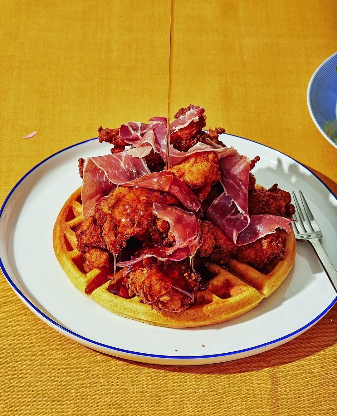 Overhead shot of fried chicken and waffle dish with prosciutto and syrup on a white plate against a yellow tablecloth background.