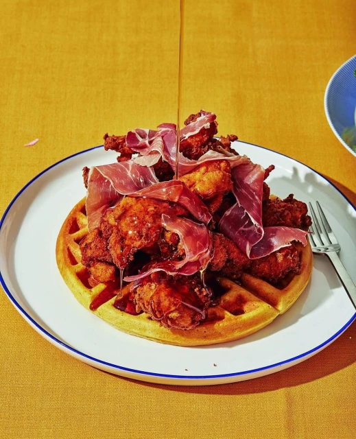 Overhead shot of fried chicken and waffle dish with prosciutto and syrup on a white plate against a yellow tablecloth background.