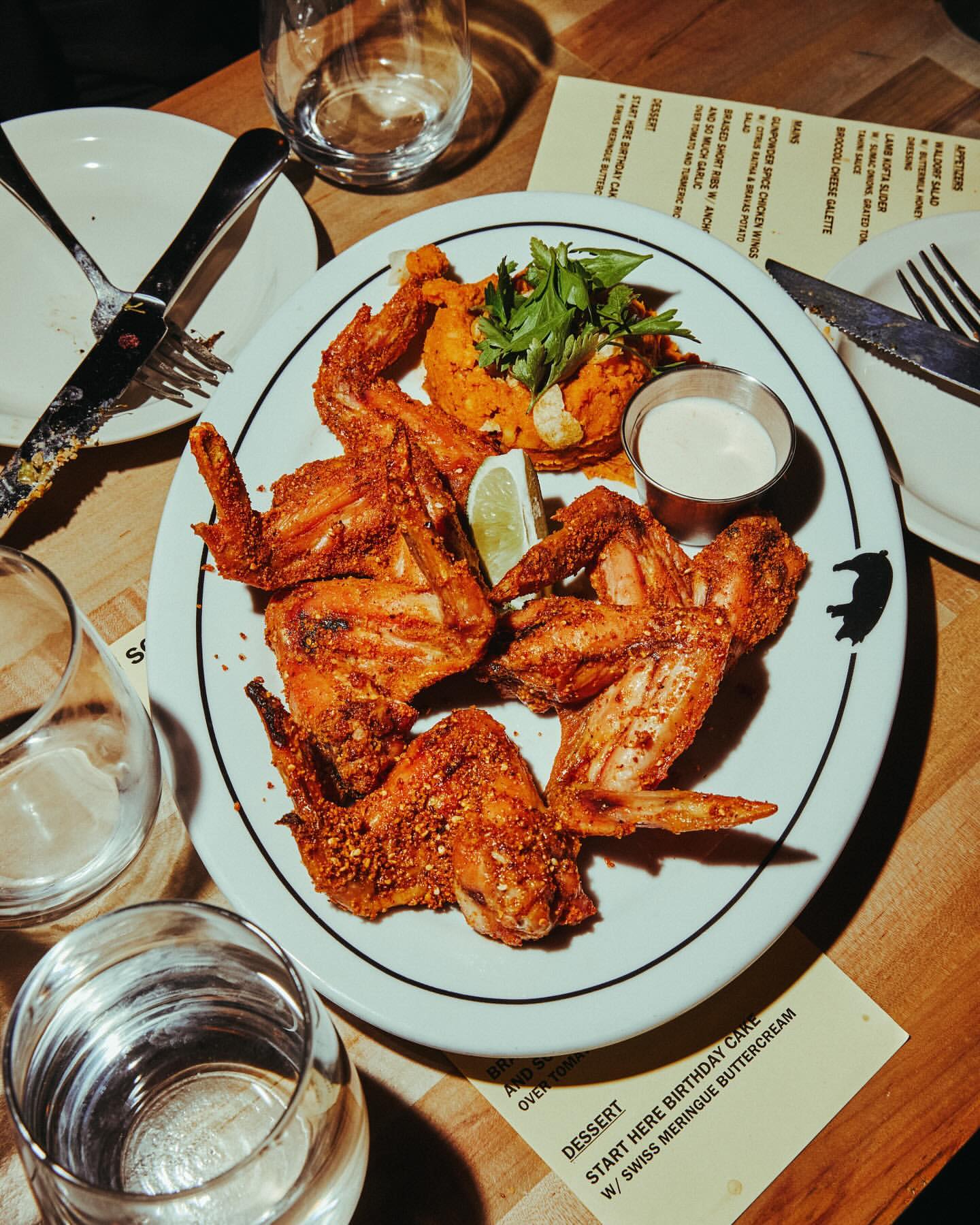 Overhead shot of fried chicken wings served on a plate with a side of sweet potato and cream sauce, complemented by drinks and cutlery.