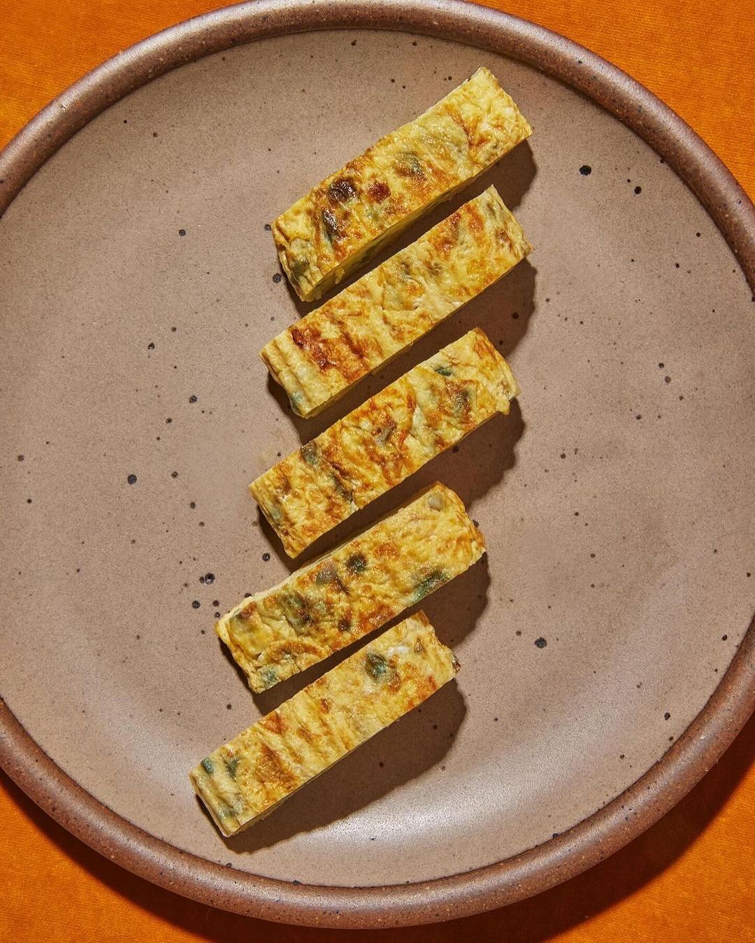 Overhead shot of five sliced tamagoyaki omelets arranged on a speckled ceramic plate, set against an orange backdrop. Food photography.