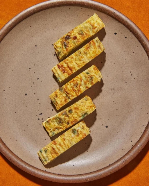 Overhead shot of five sliced tamagoyaki omelets arranged on a speckled ceramic plate, set against an orange backdrop. Food photography.