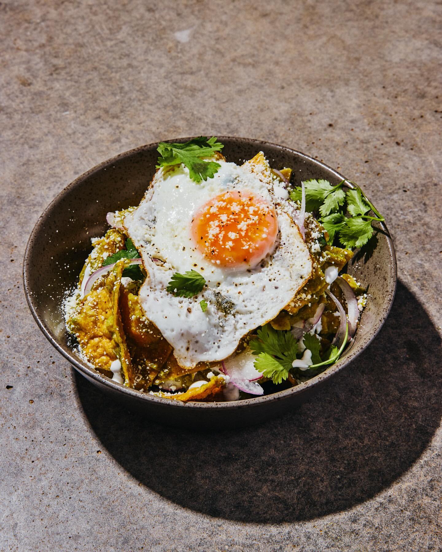 Overhead shot of Chilaquiles dish with fried egg, tortilla chips, salsa verde, onion, cilantro in a dark bowl on a textured surface.