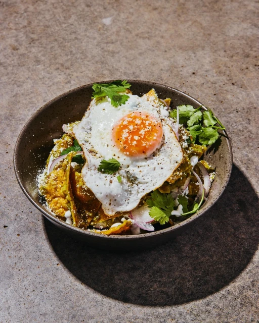 Overhead shot of Chilaquiles dish with fried egg, tortilla chips, salsa verde, onion, cilantro in a dark bowl on a textured surface.