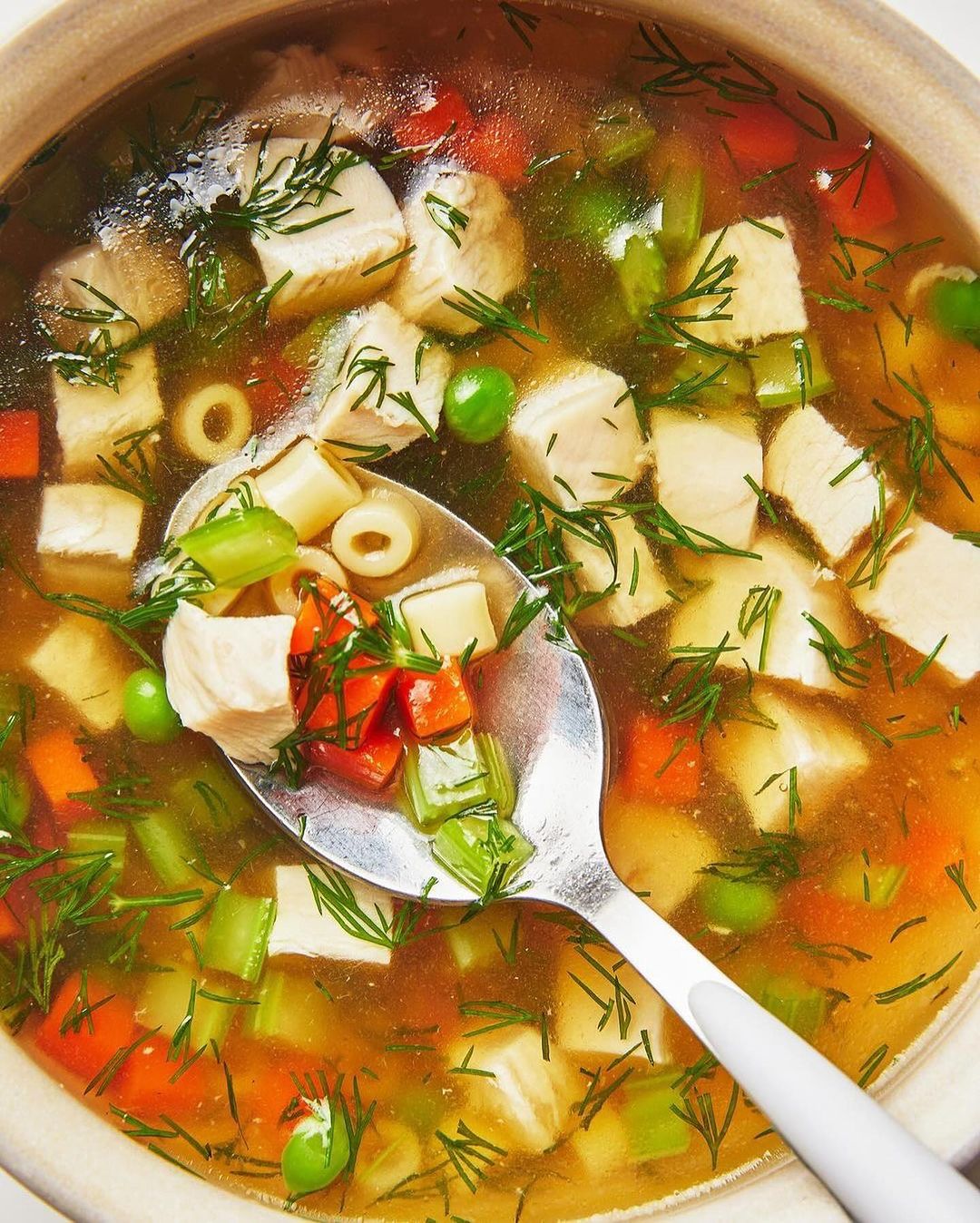 Overhead view of a savory chicken and vegetable soup with ditalini pasta in a ceramic bowl, garnished with dill, with a silver spoon.