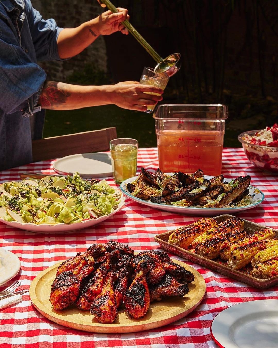 Outdoor barbecue with person pouring drink, table with grilled chicken, corn, salad, and a red-and-white checkered tablecloth.