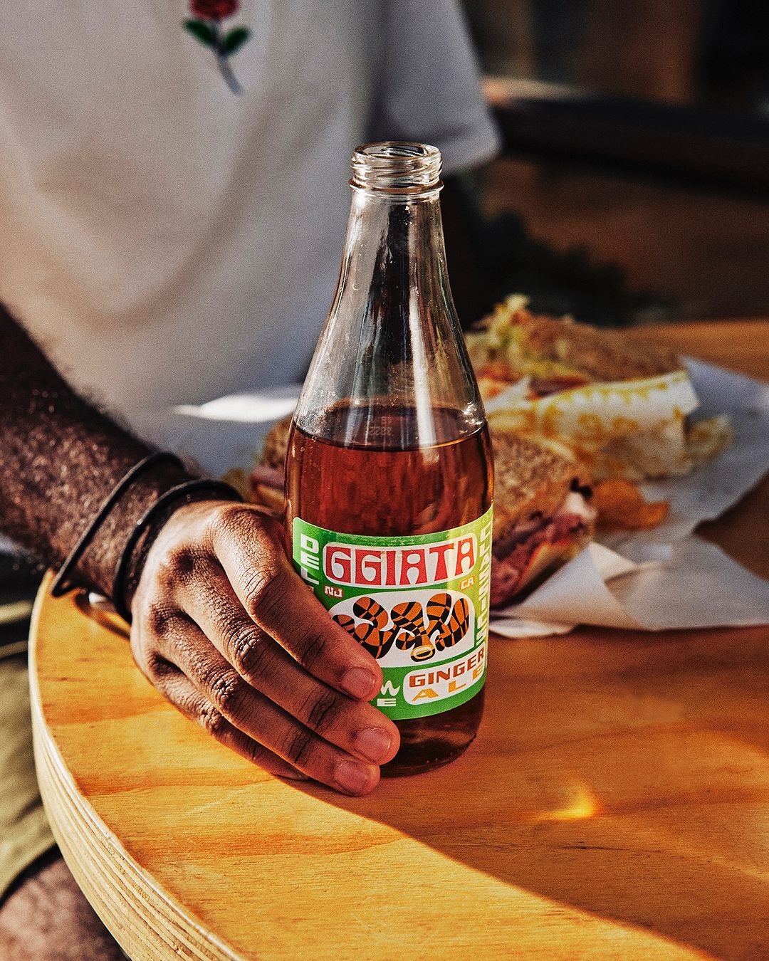 A person holds a bottle of ginger ale at a table, with a sandwich and drink next to them in a naturally lit, outdoor setting.