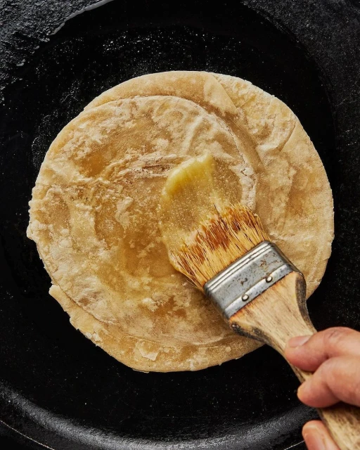 Overhead shot of a person brushing clarified butter, or ghee, onto a round flatbread cooking on a black pan for Indian cuisine.