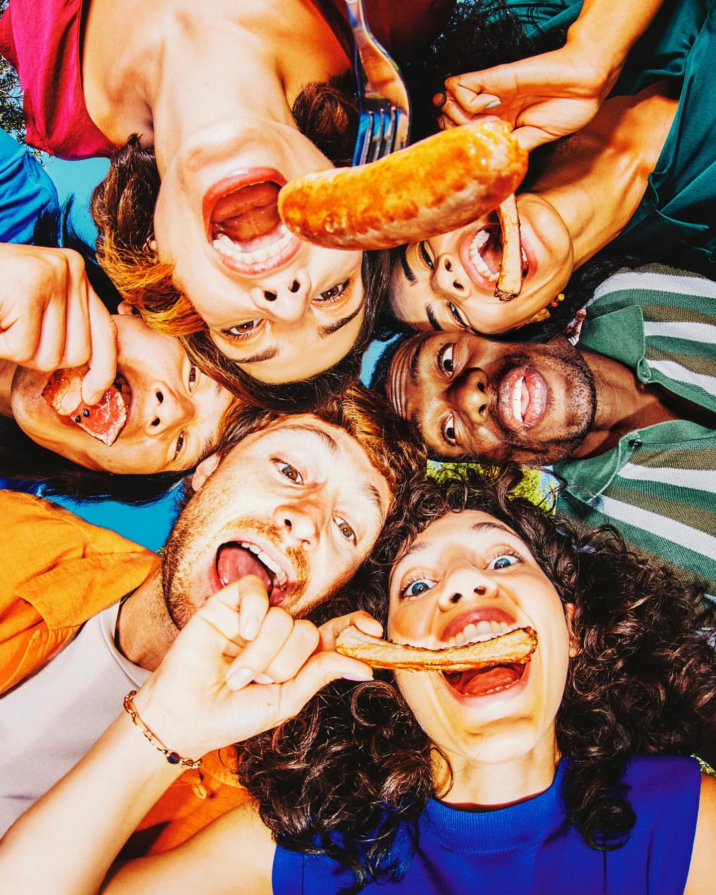 Group of diverse young adults outdoors, smiling and eating sausage, captured in a high-angle, close-up shot with natural lighting.