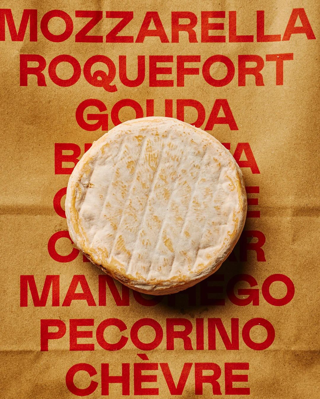 Overhead shot of round soft cheese on brown paper printed with various cheese names in red, showcasing a food photography composition.