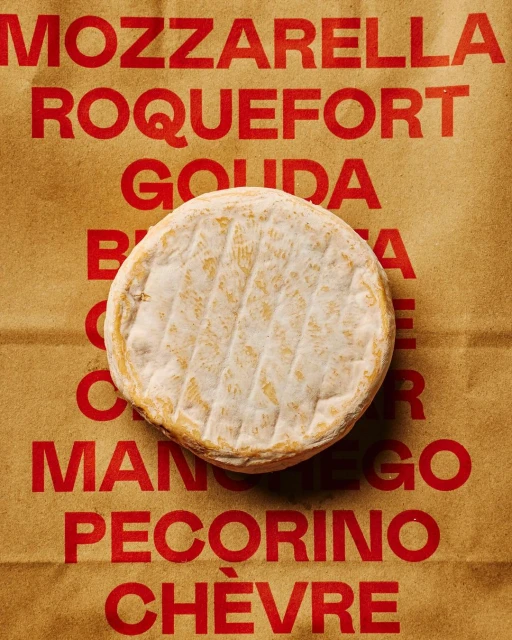 Overhead shot of round soft cheese on brown paper printed with various cheese names in red, showcasing a food photography composition.