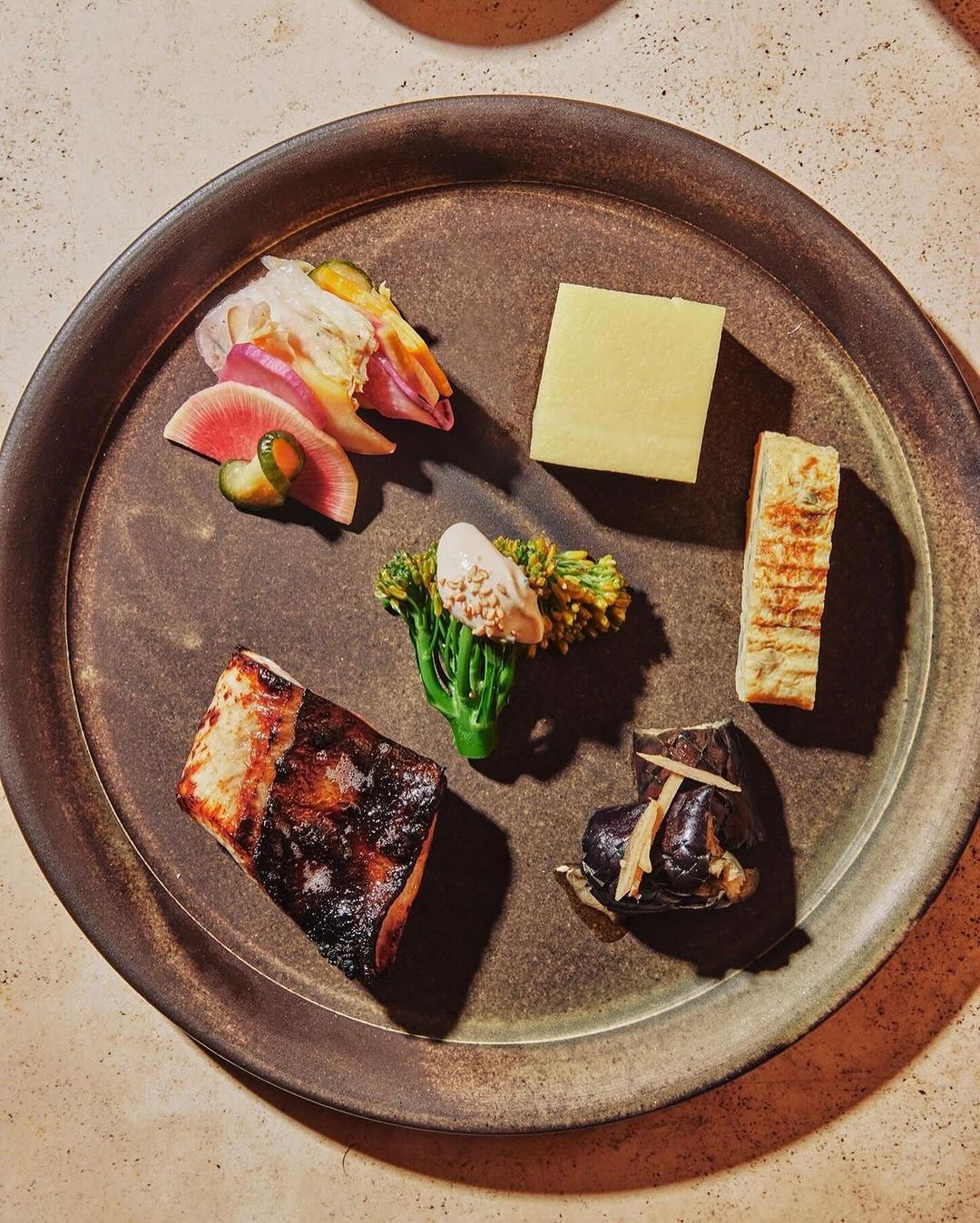 Overhead view of a dark plate with six different food preparations: grilled fish, yellow block, broccolini, sliced vegetables, and eggplant.