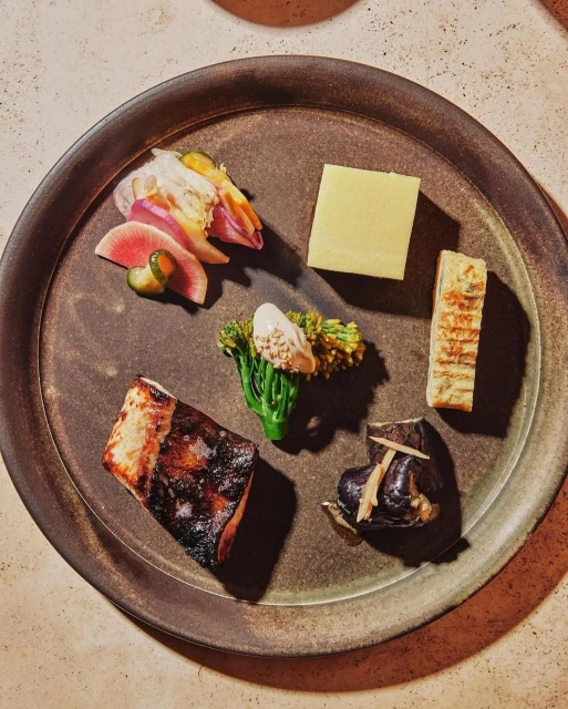Overhead view of a dark plate with six different food preparations: grilled fish, yellow block, broccolini, sliced vegetables, and eggplant.