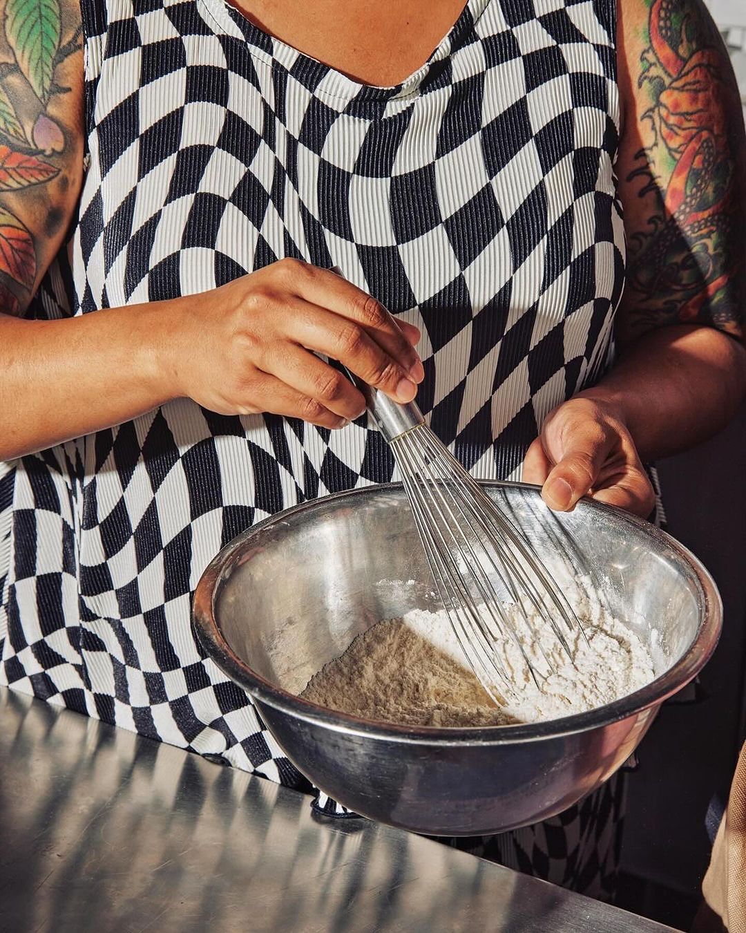 Close-up of a person with tattooed arms whisking batter in a stainless steel bowl while wearing a checkered top indoors.