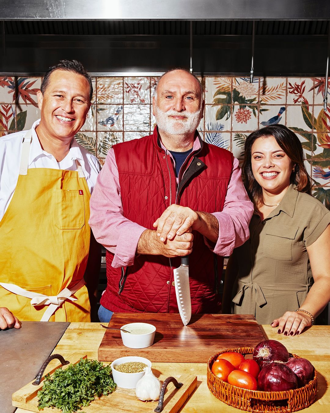 Three people in kitchen setting with food prep: an adult male in an apron, a bearded adult male with a knife, and an adult female.