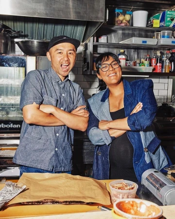 Candid portrait of two Asian chefs smiling in a commercial kitchen, with stainless steel equipment, food preparation, and food in containers.