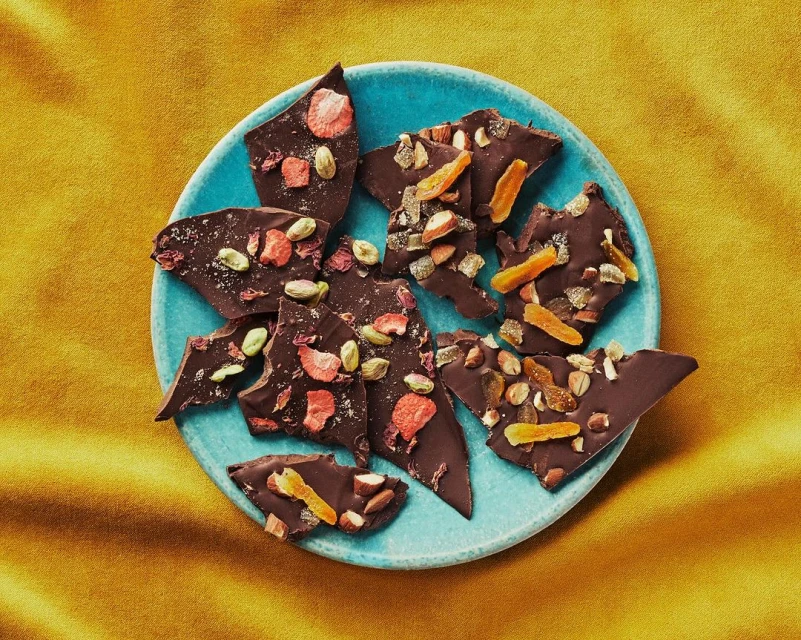 Artisan dark chocolate bark with dried fruit and pistachios on a blue plate, set against a textured yellow background.