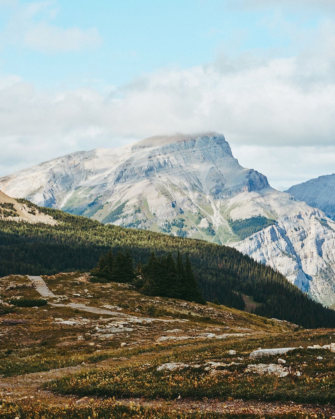 Grand mountain with cloud cover on the peak, framed by green hillside terrain and a clear sky with scattered clouds on a sunny day.