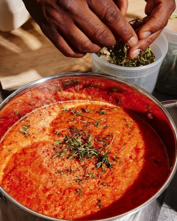 Chef prepares tomato sauce in a stainless steel pot, adding fresh thyme, indoors, with a focus on culinary preparation.