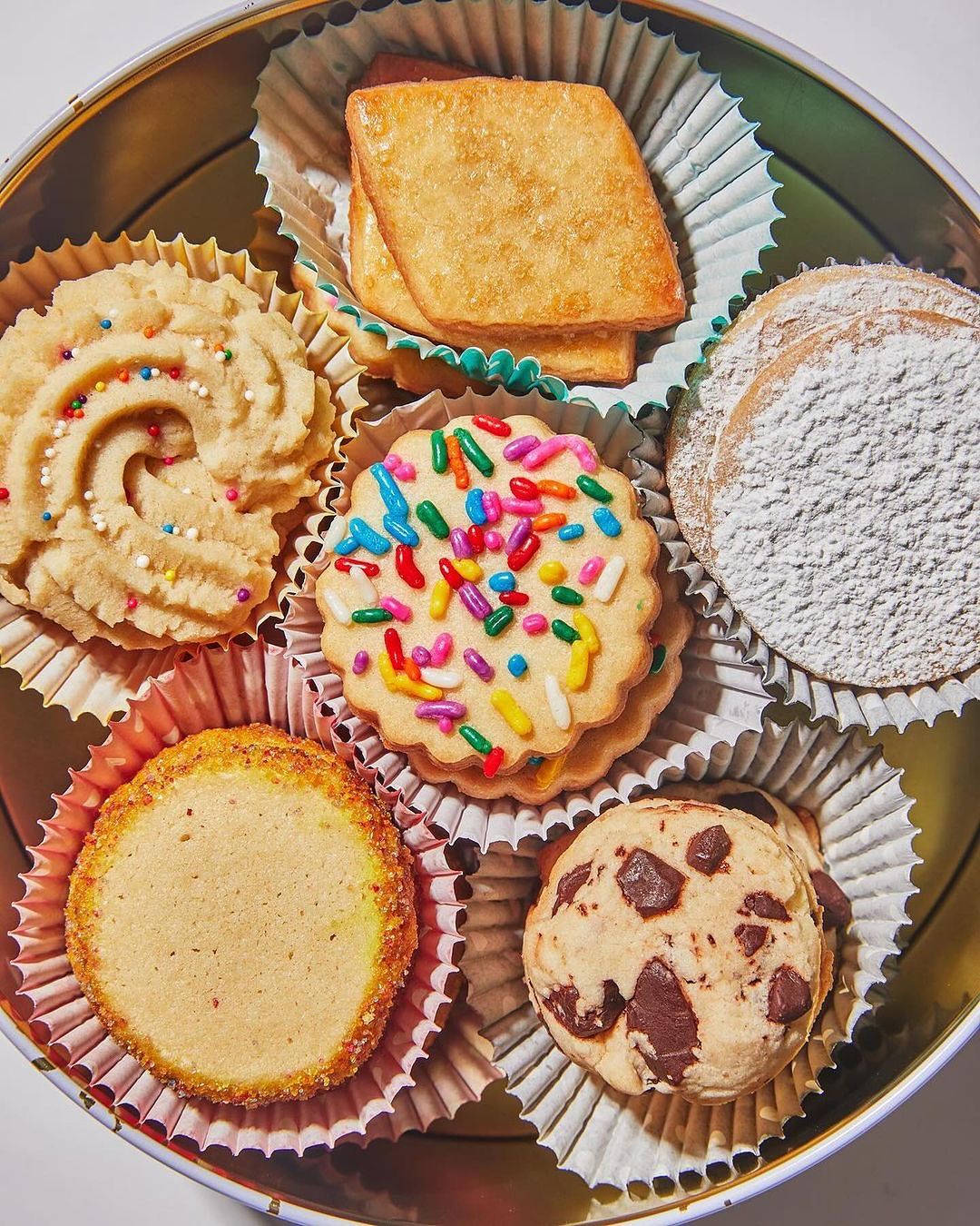 Overhead shot of an assortment of gourmet cookies in paper liners, arranged inside a round tin, suggesting a sweet treat selection.