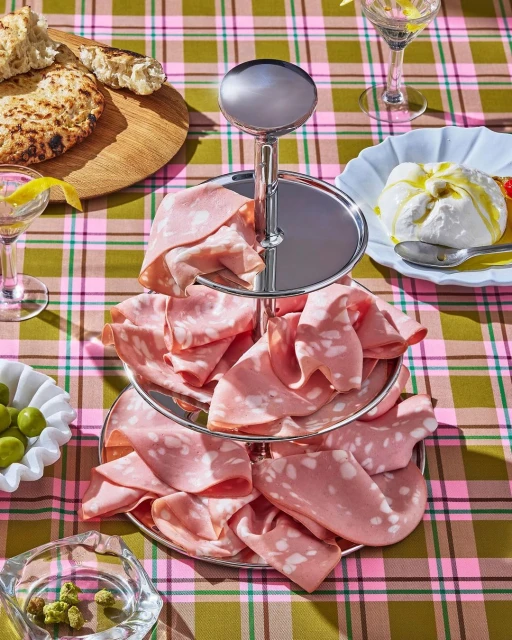 Three-tiered serving platter filled with mortadella, accompanied by bread, cheese, and olives on a pink and green plaid tablecloth.