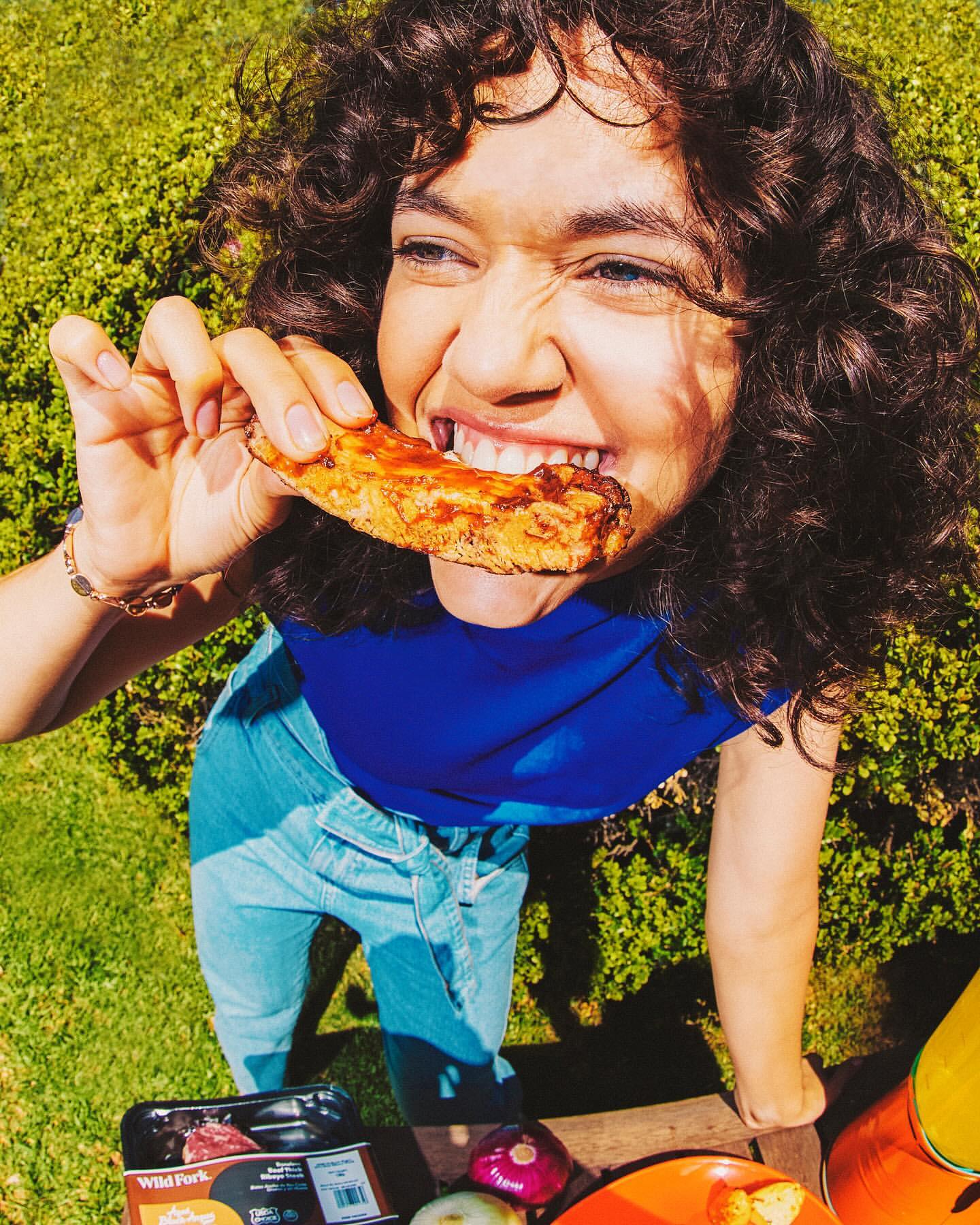 Woman with curly hair eating plant-based rib in a sunny garden. Blue shirt, jeans, plate of food, and package of ribs are visible.
