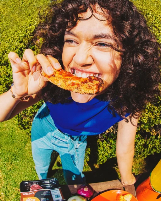 Woman with curly hair eating plant-based rib in a sunny garden. Blue shirt, jeans, plate of food, and package of ribs are visible.