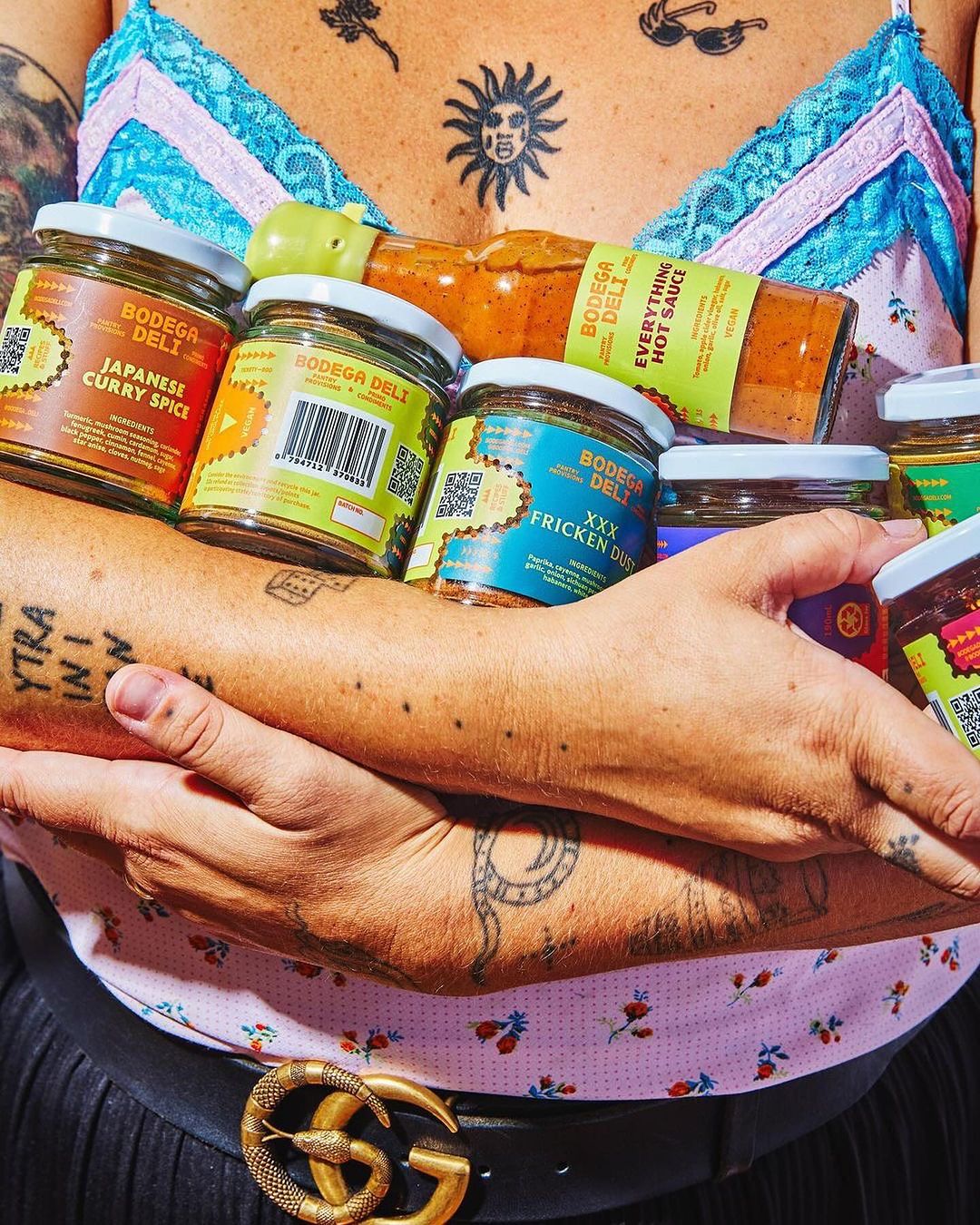 Person holding Bodega Deli hot sauce and curry spice jars in front of floral shirt and tattoos, wearing Gucci belt, in eye-level shot.