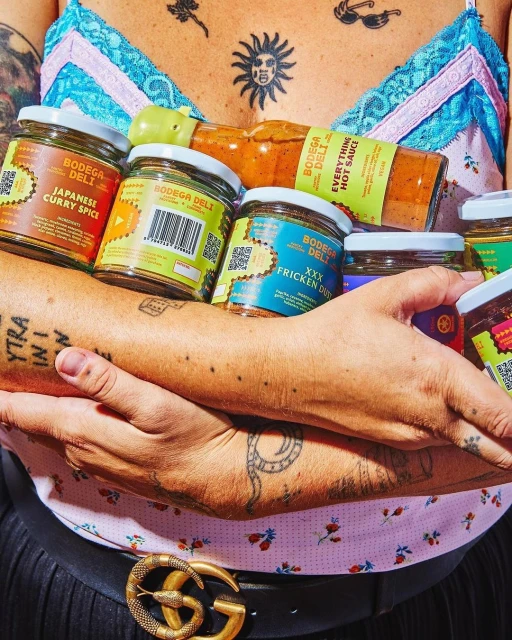 Person holding Bodega Deli hot sauce and curry spice jars in front of floral shirt and tattoos, wearing Gucci belt, in eye-level shot.