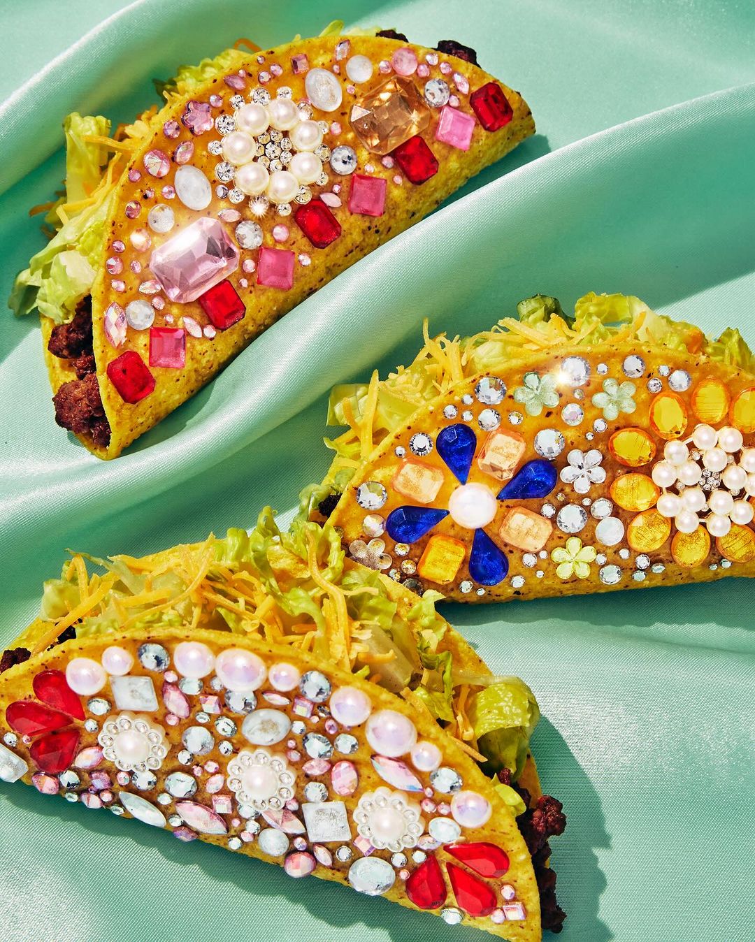 Overhead shot of three tacos embellished with colorful jewels, pearls, and gems, placed on a mint green silk fabric background.