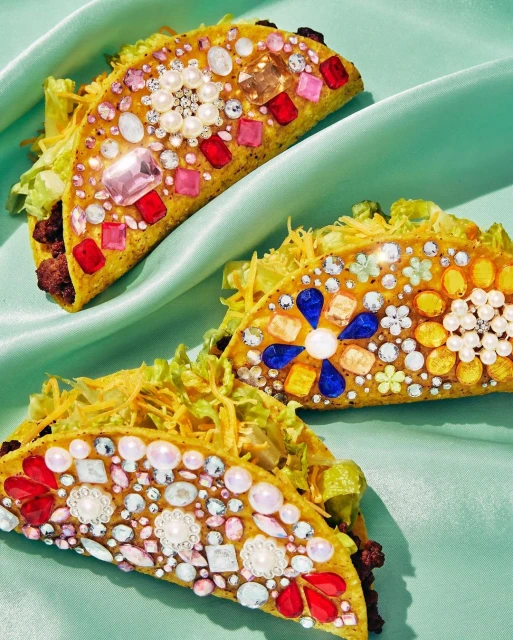 Overhead shot of three tacos embellished with colorful jewels, pearls, and gems, placed on a mint green silk fabric background.