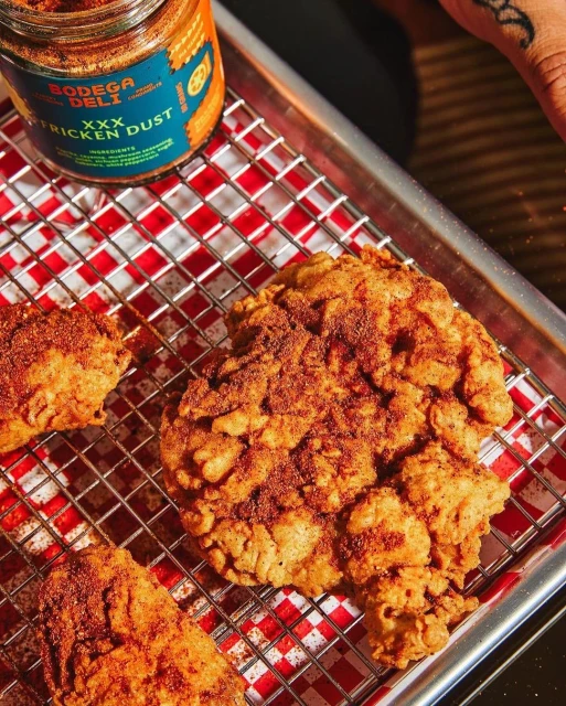 Golden fried chicken pieces sit on a wire rack over a red and white checkered surface, with a jar of spice in the background.