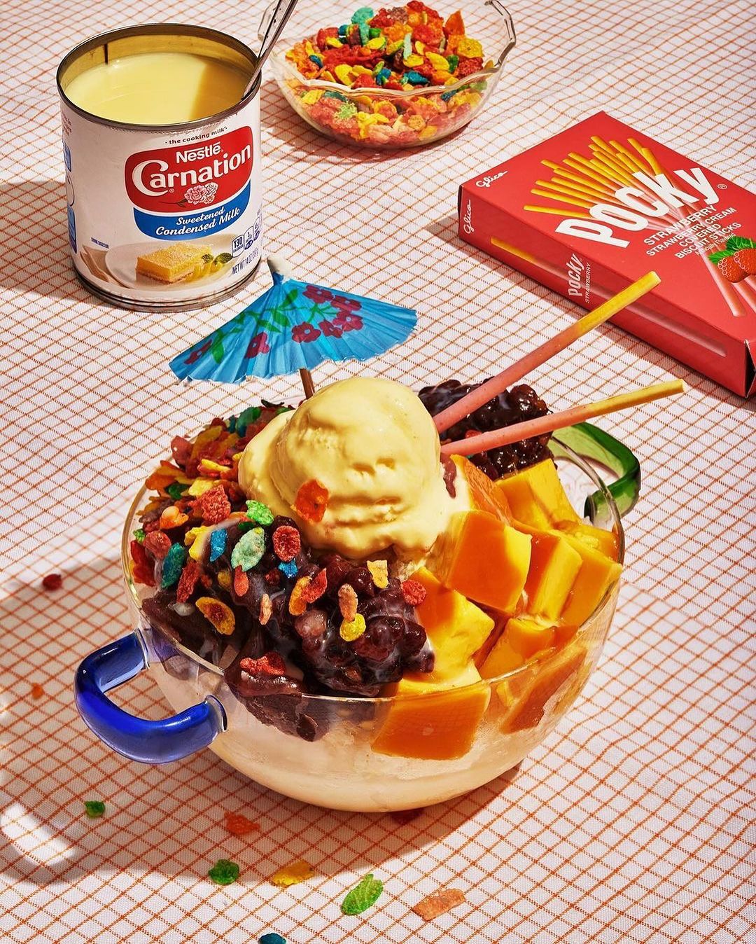 Halo-halo dessert in a glass bowl, featuring ice cream, mango, red beans, fruit loops, topped with a parasol, on a checkered surface.