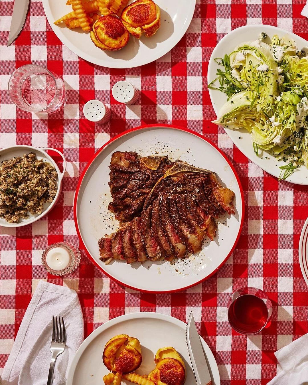 Overhead shot of a steak dinner on a red-and-white checkered tablecloth, featuring a steak, salad, potatoes, and a glass of red wine.