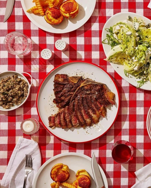 Overhead shot of a steak dinner on a red-and-white checkered tablecloth, featuring a steak, salad, potatoes, and a glass of red wine.