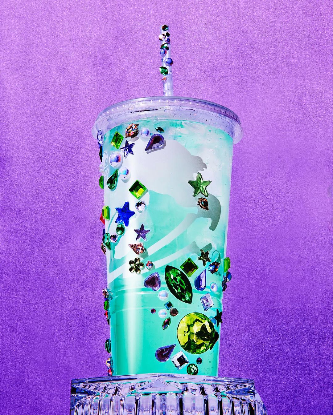 A clear plastic tumbler with a lid and straw, decorated with various gems and stones, sits on a clear glass base against a purple background.