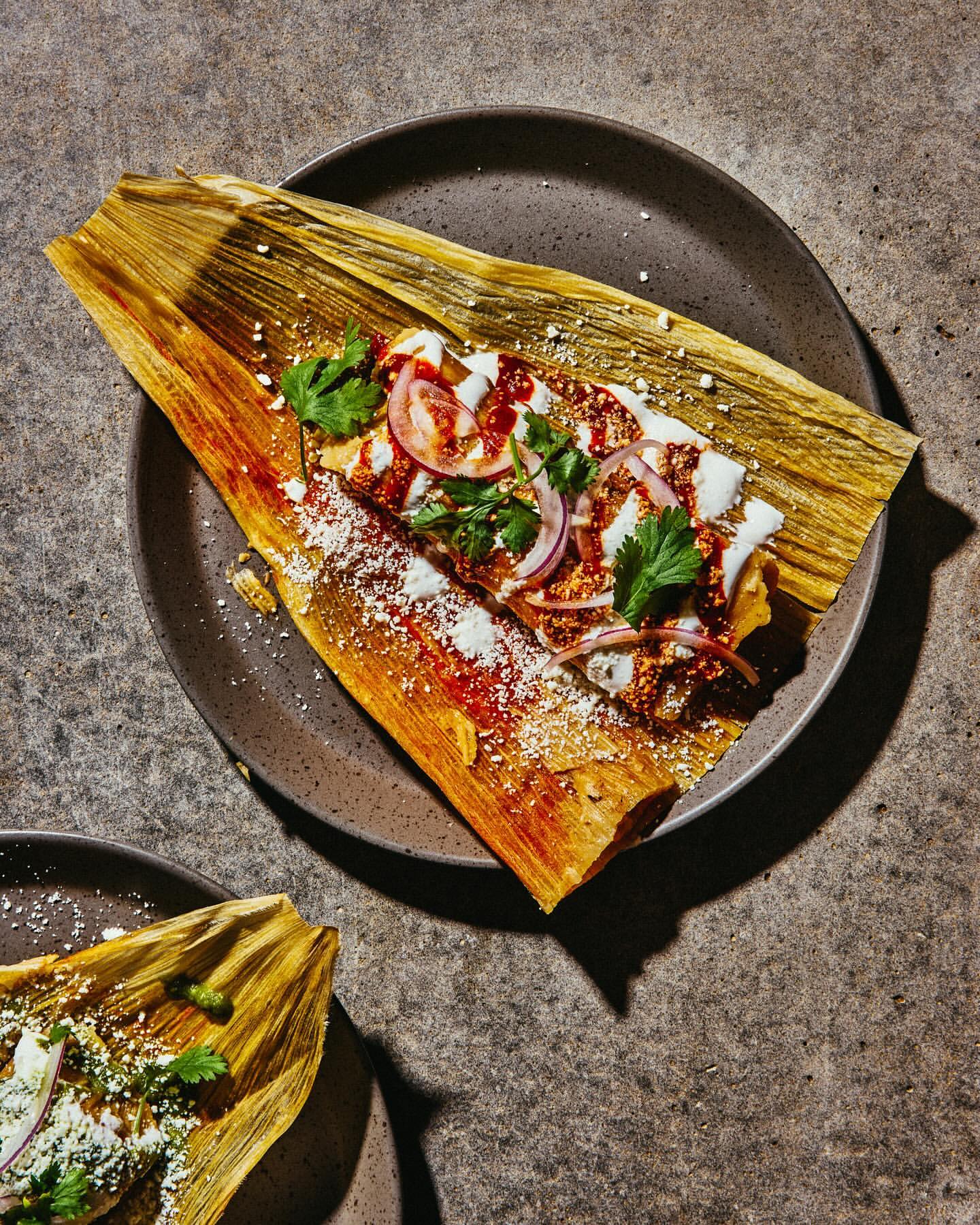 Overhead shot of a beautifully plated tamale on a gray plate, garnished with onions, cilantro, cheese, and sauce, atop a textured surface.